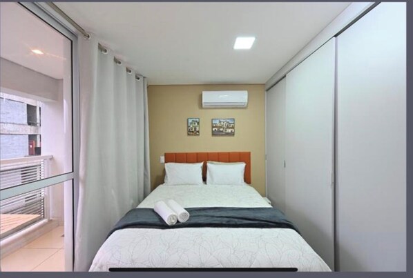 Habitació d'hotel amb un llit, una finestra amb cortines, un aire condicionat i dues imatges penjades a la paret.