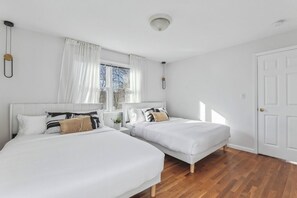 Et soveværelse med to senge, et vindue med gardiner, en hvid dør og en lampe i loftet.