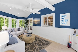 A modern living room with a white ceiling fan, a large window with a view of greenery, a white sofa, a round coffee table, and a 'meet me at the boat' sign on the wall. - Gorgeous, Stylish Home - Ping Pong & Firepit (Chatham)
