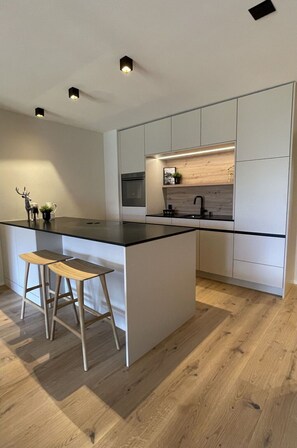 Eine moderne Küche mit einer zentralen Kochinsel, Holzstühlen, einem Einbauofen und einer Holzrückwand.