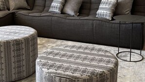 Ein modernes Wohnzimmer mit einem grauen Sofa, gemusterten Hocker und zwei gerahmten Fotografien an der Wand.