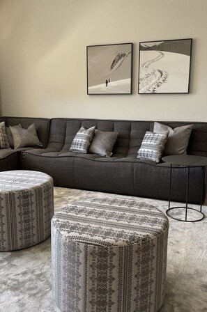 Ein modernes Wohnzimmer mit einem grauen Sofa, gemusterten Hocker und zwei gerahmten Fotografien an der Wand.
