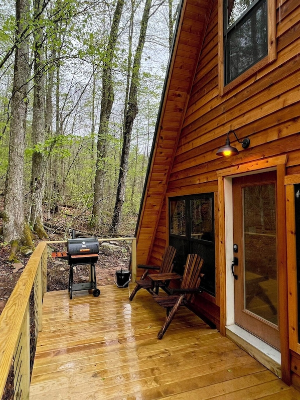 Una terraza de madera con una parrilla, sillas, un cerramiento de vidrio y un fondo de bosque.