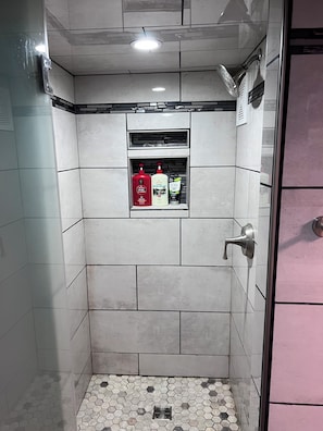 Et badeværelse med hvide fliser, en bruser, en glasdør og en hylde med toiletartikler.