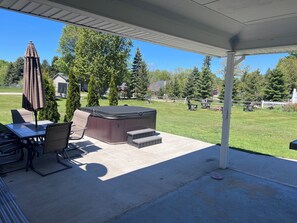 Un patio couvert avec un jacuzzi, des sièges d’extérieur et une vue sur une pelouse verdoyante, des arbres et une clôture blanche en bois.