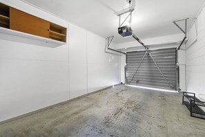 A spacious garage with a closed metal garage door, a ceiling-mounted projector, and a wooden shelf. - Warmth and the spacious 2 double bedroom (Manurewa)