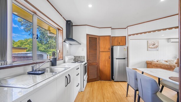 Een kleine keuken met een spoelbak, fornuis en koelkast, houten keukenkastjes, en een eethoek met een tafel en stoelen.
