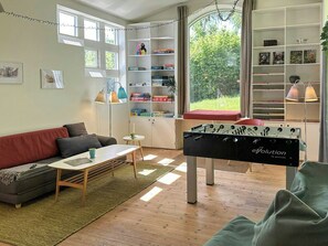 Un salón acogedor con sofá, una mesa de centro y una mesa de futbolín. Hay estanterías con libros y objetos decorativos, y una ventana con vistas al verdor.