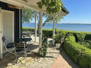 Un patio cubierto con mesa y sillas, vista al mar y un seto.