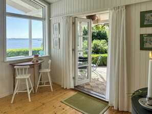 Una habitación con suelo de madera, una mesa con sillas, un ventanal con vista al mar y una puerta que da al exterior.
