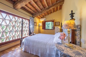 Ein Schlafzimmer mit Holzboden, einem Bett mit weißer Bettdecke, einem Fenster mit Gitter und einem Nachttisch mit Lampe und Blumen.