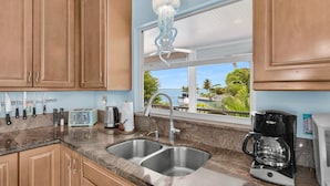 Una cocina con armarios de madera, un lavamanos doble, una cafetera y vistas a un paisaje tropical.