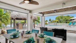 Un área de comedor con vista a la piscina, mobiliario exterior y una estructura de techo de paja.