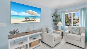 Una sala de estar con un televisor mostrando una escena de playa, dos sillones blancos, una mesita con una lámpara y una planta.