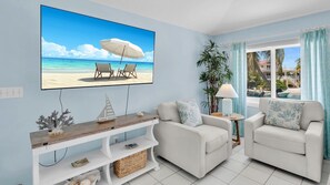Ein Wohnzimmer mit Fernseher, der eine Strandansicht zeigt, zwei weißen Sesseln, einem kleinen Tisch mit Lampe und einer Pflanze.