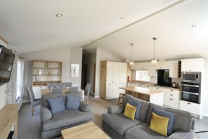 A modern living room with a grey sofa, a wooden coffee table, and a TV on the left. The kitchen area is visible with white cabinets, a microwave, and a stove.