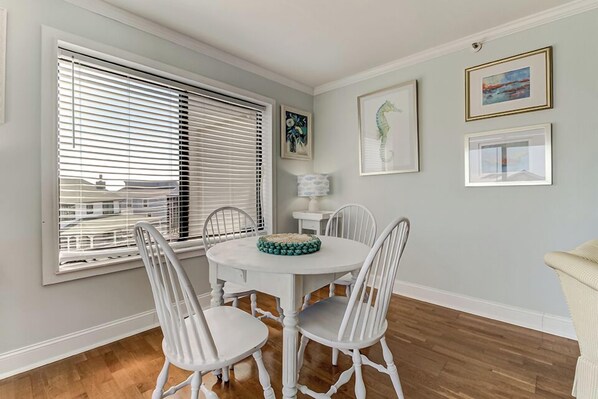 A dining area with a round table, four chairs, a window with blinds, and framed artwork on the wall. - Ocean Views, Balcony, Pool, STEPS to Beach Restaurants Shops (Wrightsville Beach)