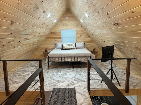 Una habitación con techo alto de madera, una cama, una mesita y un ventanal.