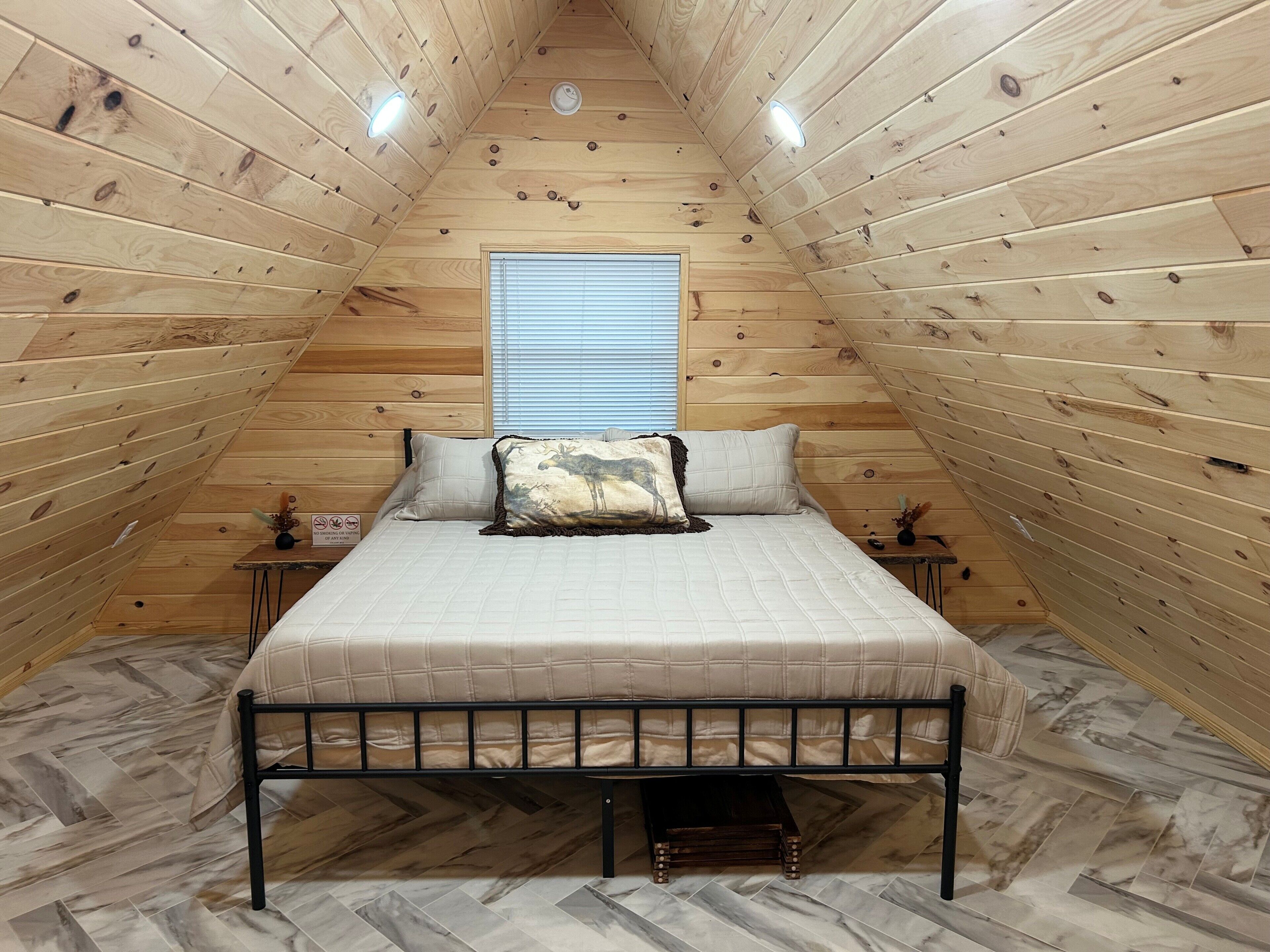 Dormitorio tipo loft con techo inclinado de madera, una cama con cabecera, dos mesitas de noche y una ventana con persianas.