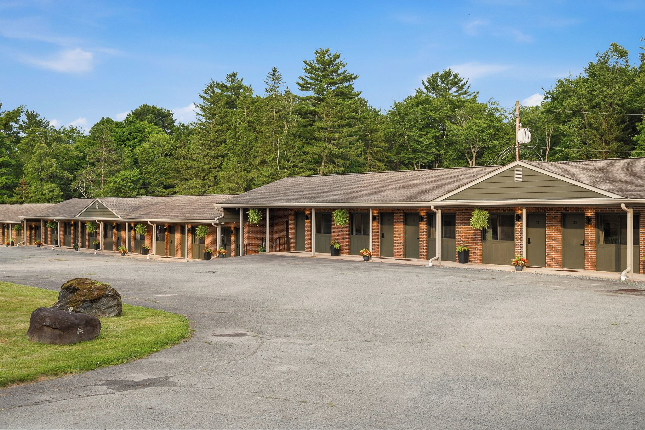 A row of motel buildings with individual rooms, a parking lot, and a forested backdrop.