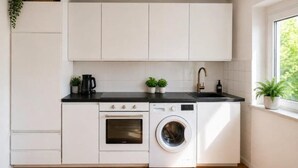 Une cuisine compacte avec des meubles blancs, un plan de travail noir, un four encastré, un lave-linge et une fenêtre donnant sur un beau paysage.