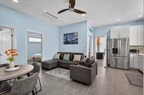 A modern living room with a sectional sofa, a dining table with chairs, a kitchen with stainless steel appliances, and a ceiling fan.