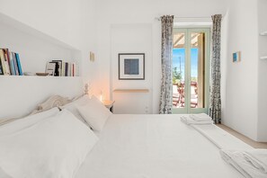 Ein Schlafzimmer mit Bett, Nachttisch, Lampe, Bücherregal, gerahmtem Bild und Blick durch eine Tür.