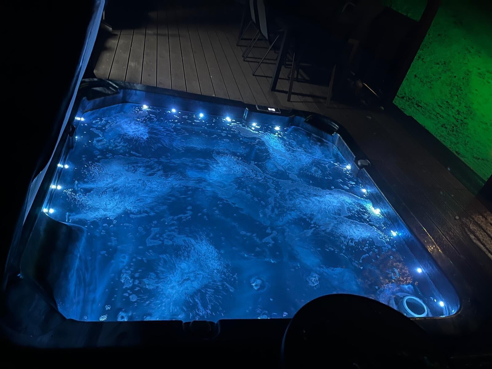 A hot tub with lights, surrounded by a wooden deck and a green wall.