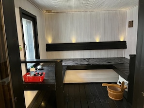 A sauna room with a bench, a wooden bucket, and a red basket with toiletries.