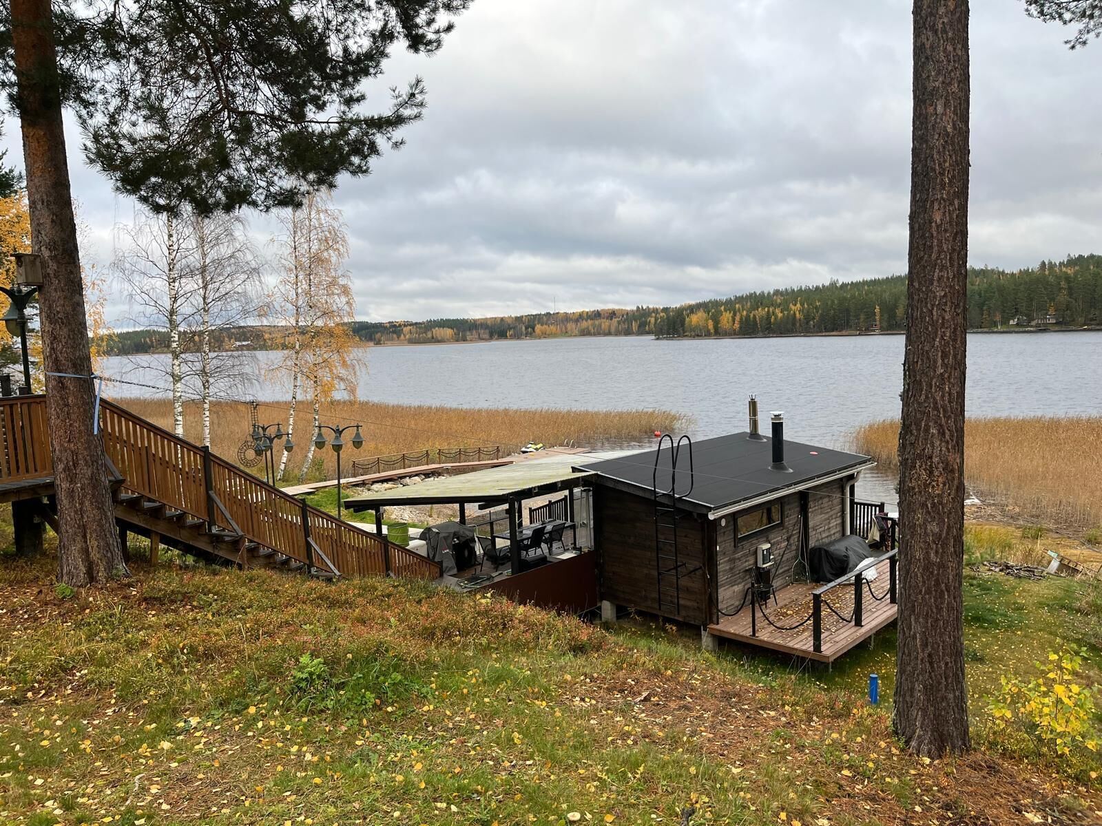 A lakeside cabin with a deck, a small building with a chimney, and a wooden staircase leading to a platform.