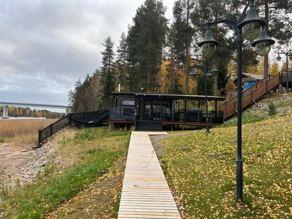 A wooden pathway leading to a modern building with a glass facade, surrounded by trees and a distant bridge.