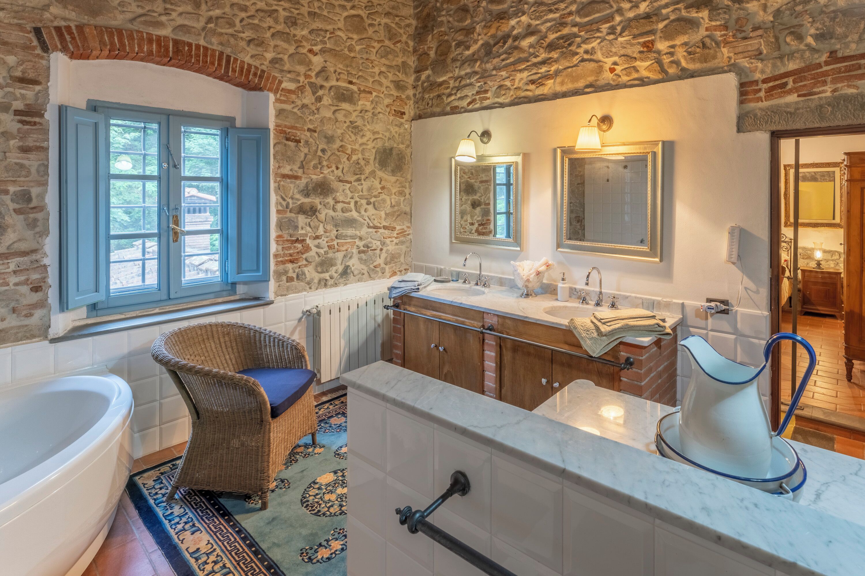 Une salle de bain avec un mur en pierre, une baignoire, un fauteuil en osier, un lavabo et un miroir.