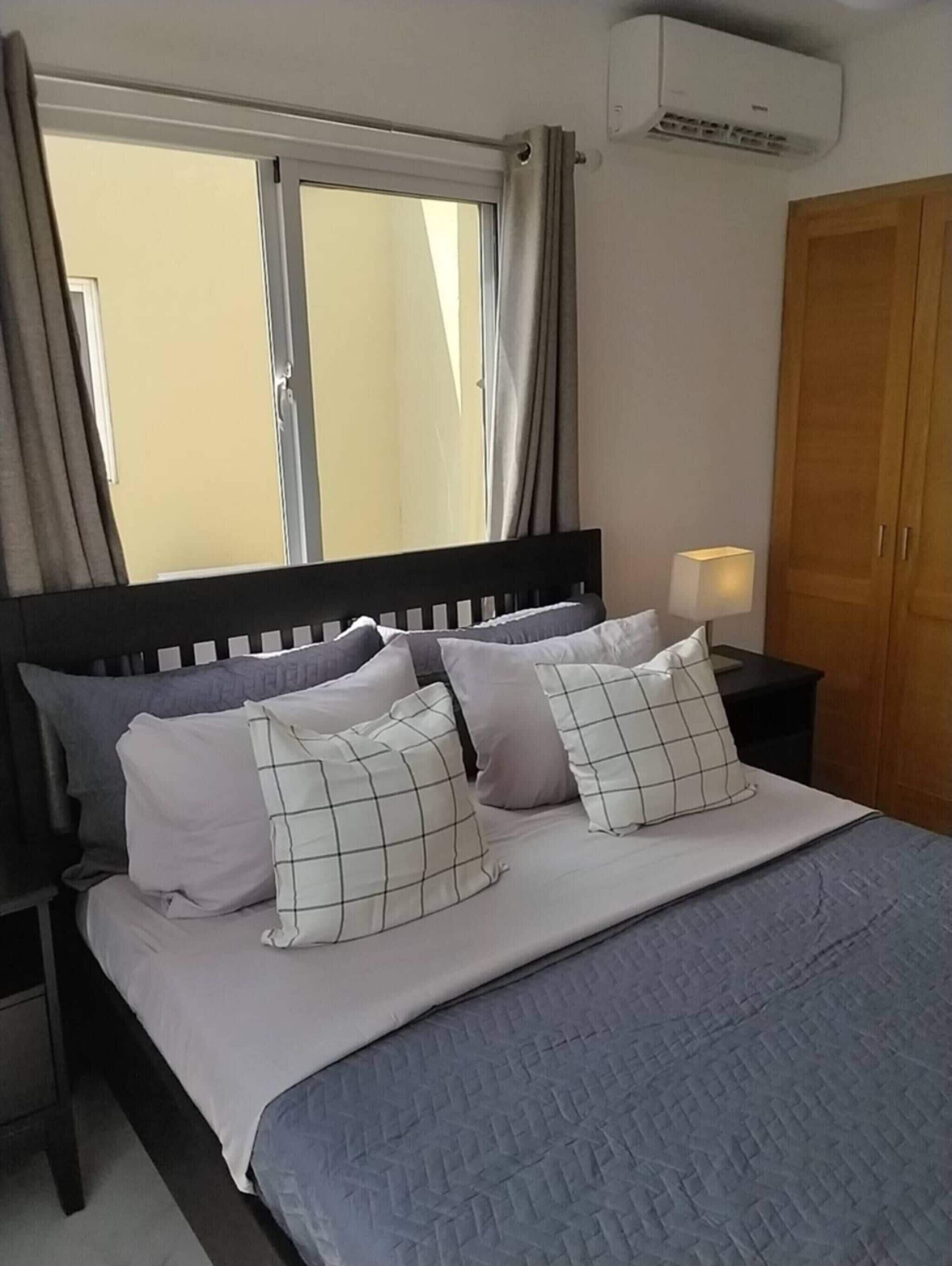 A bedroom with a bed, pillows, a nightstand, an air conditioner, and a wardrobe.