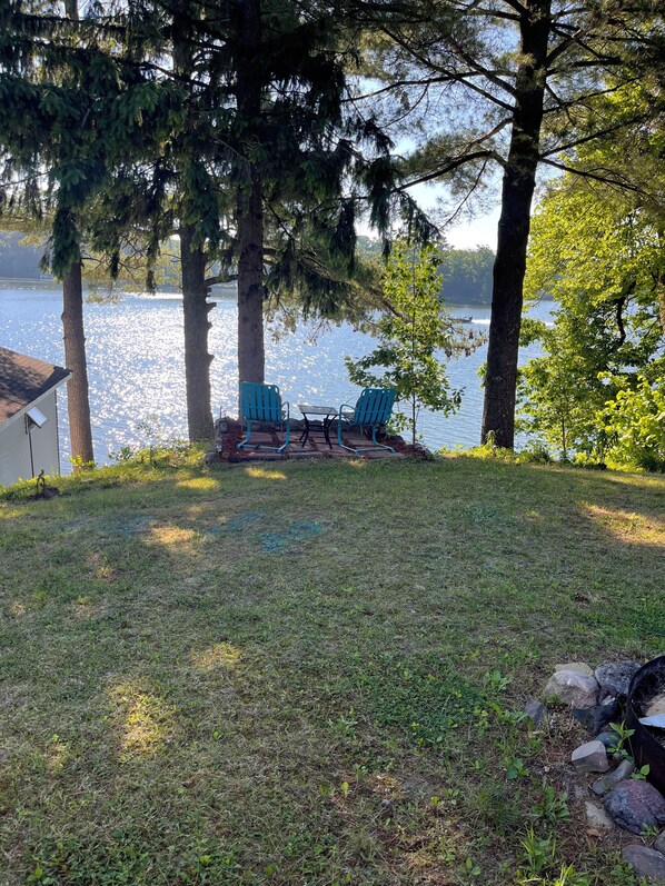 A grassy area with two blue chairs and a small table, overlooking a lake with trees and a building in the background.