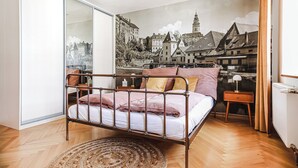 A bedroom with a metal bed frame, a large window, a cityscape mural, a wooden floor, and a small bedside table with a lamp.