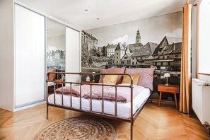 A bedroom with a metal bed frame, a large window, a cityscape mural, a wooden floor, and a small bedside table with a lamp. - Apartments Masaryk (Ceský Krumlov)