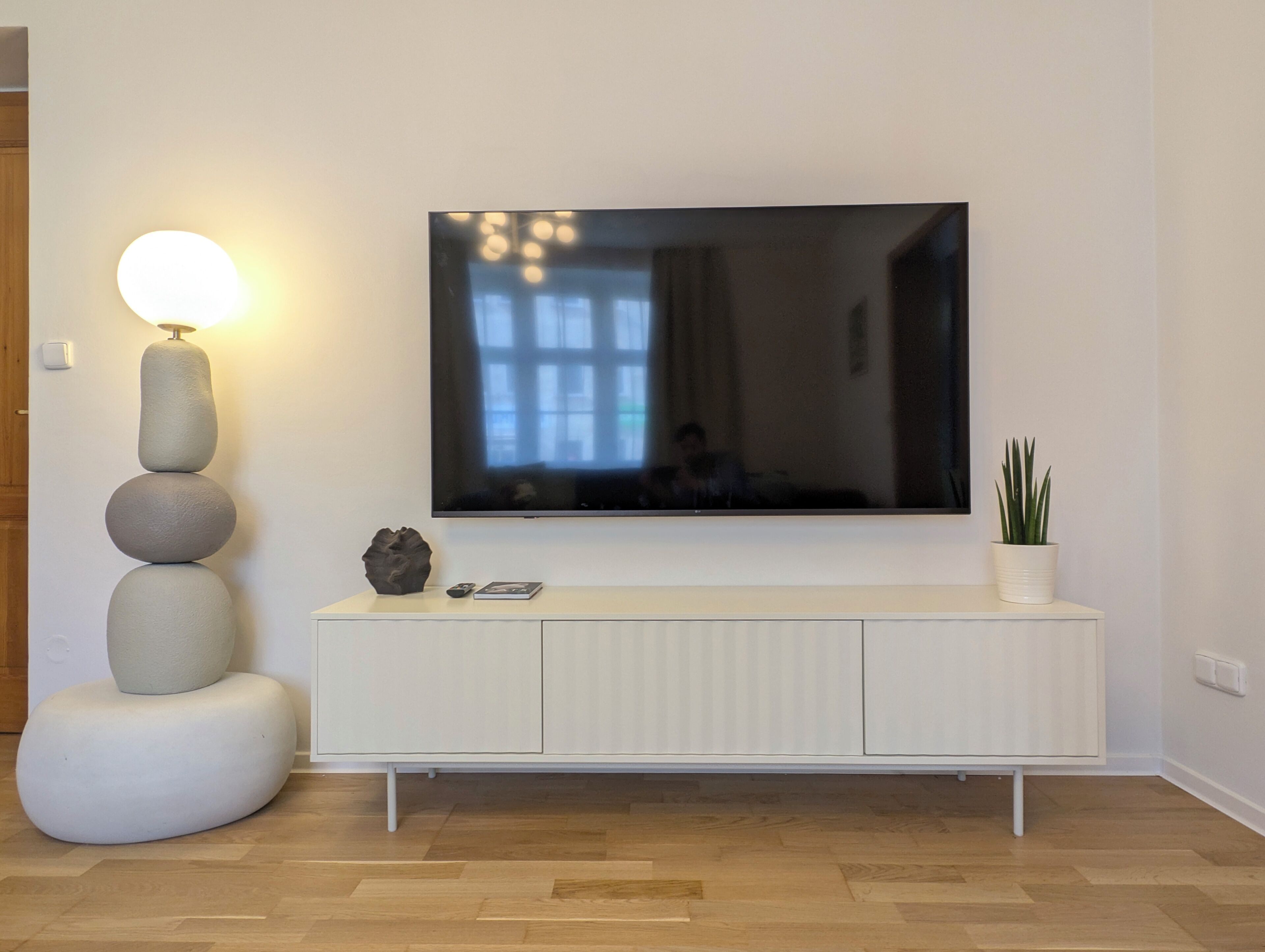 A modern living room with a large flat-screen TV mounted on a white console, a tall floor lamp, and a potted plant.