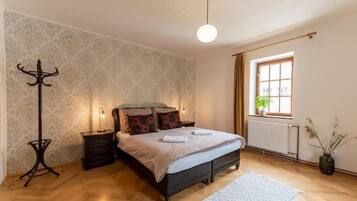 Un dormitorio con cama, mesitas de noche, una lámpara de pie, ventana con cortinas y un radiador.