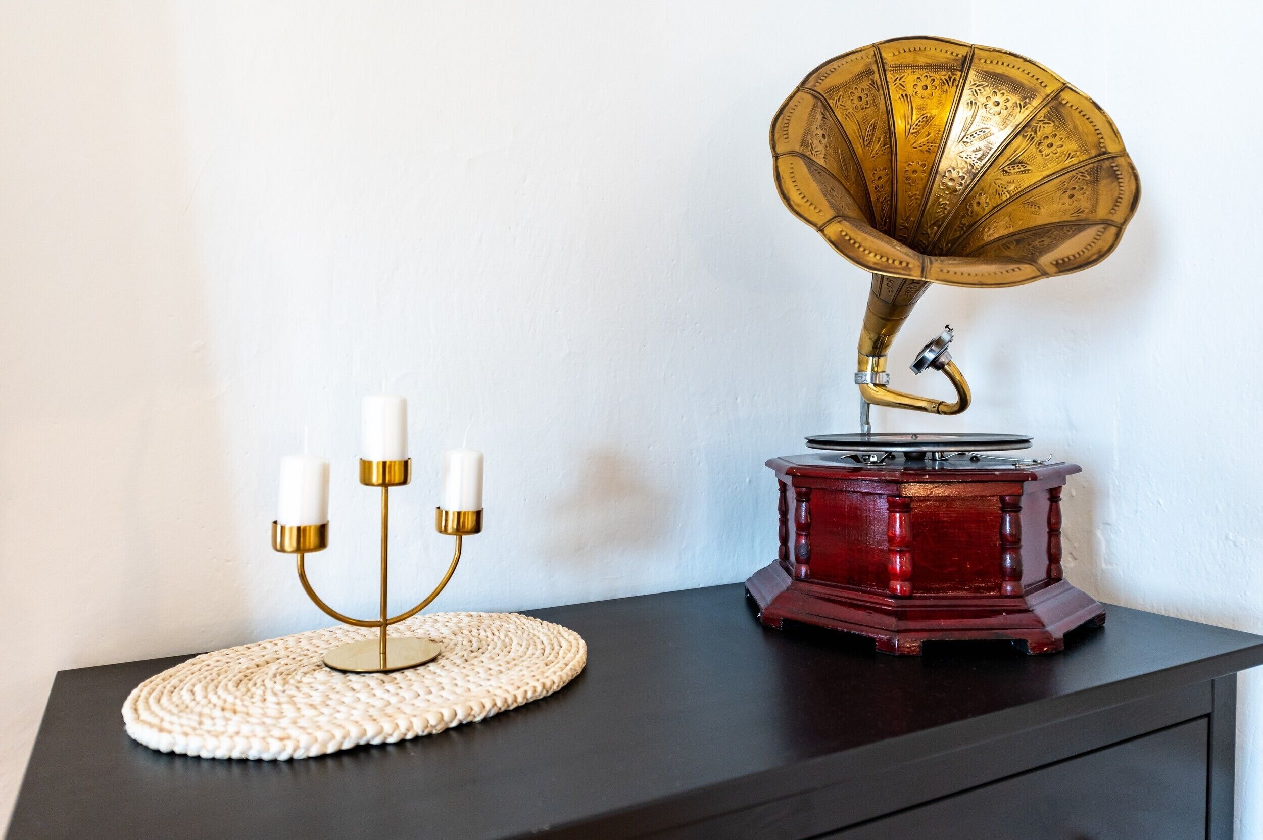 A gramophone on a dark surface with a woven mat, a candelabra with candles, and a plain wall.