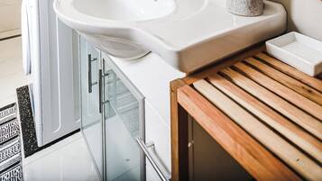 Un baño con lavamanos blanco, espejo, una pila de toallas y zona de ducha.