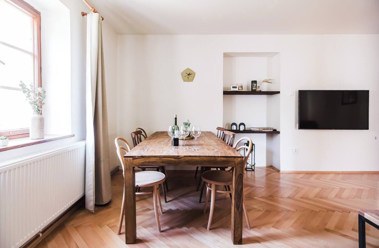 Une salle à manger avec une table en bois, six chaises, une horloge au mur, un téléviseur fixé au mur et une fenêtre avec des rideaux.