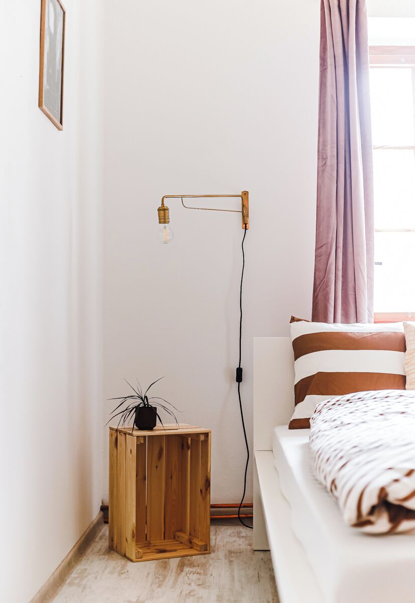 Un dormitorio con una cama, una mesita de noche con una planta, una lámpara de pie y un ventanal con cortinas.