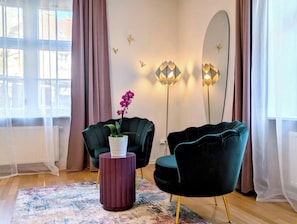 Una sala de estar moderna con dos sillones verde oscuro, una mesita redonda con base morada, una orquídea en maceta y apliques de pared decorativos.