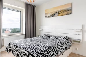 A bedroom with a bed, a window with curtains, a view of buildings, and a painting on the wall.