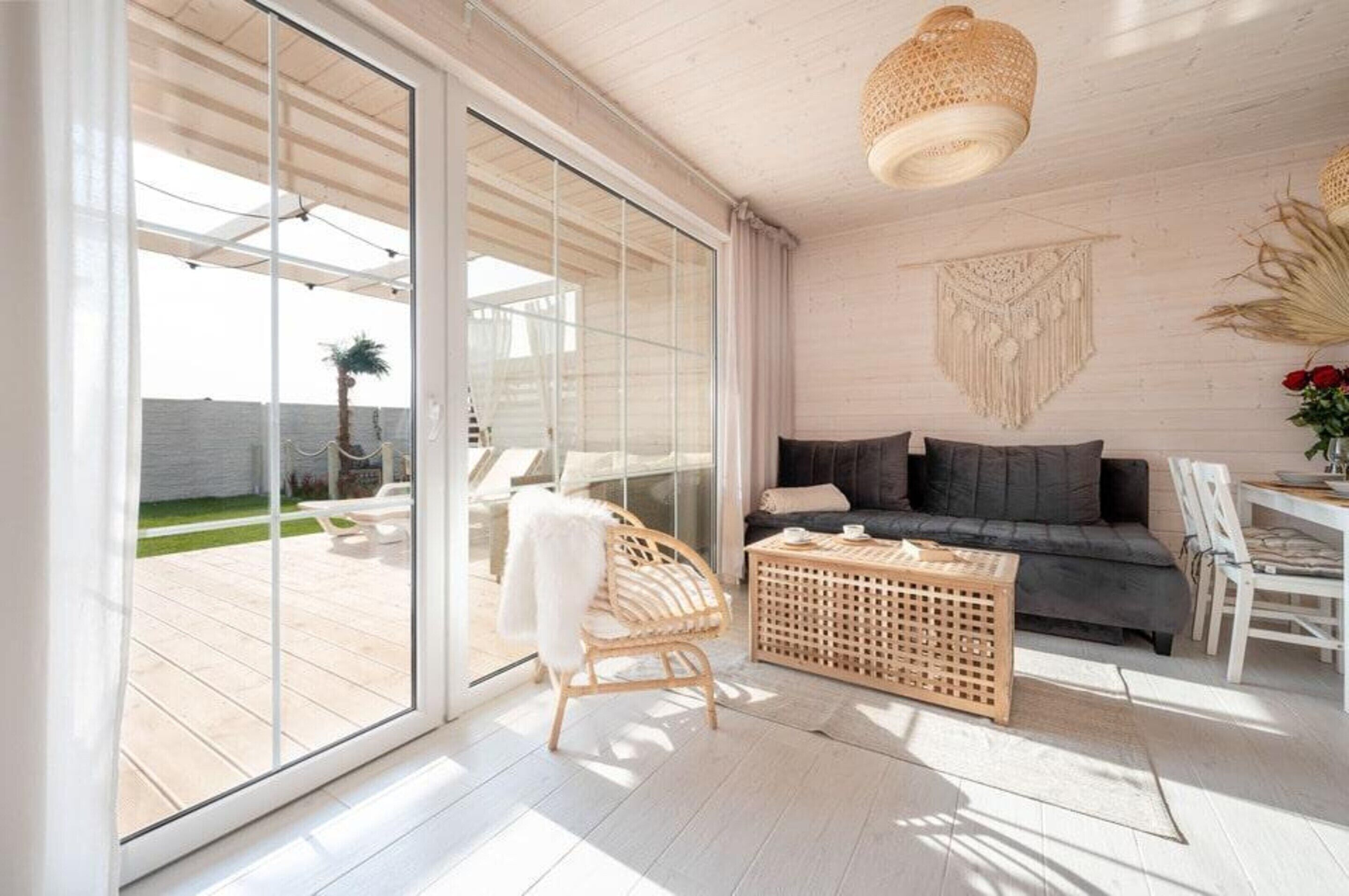 Een moderne woonkamer met een bank, een rieten stoel, een houten bijzettafeltje en uitzicht door grote glazen deuren op een terras.