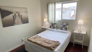 Un dormitorio con una cama, dos mesitas de noche con lámparas, una ventana con cortinas, un cuadro en la pared y vistas de edificios.