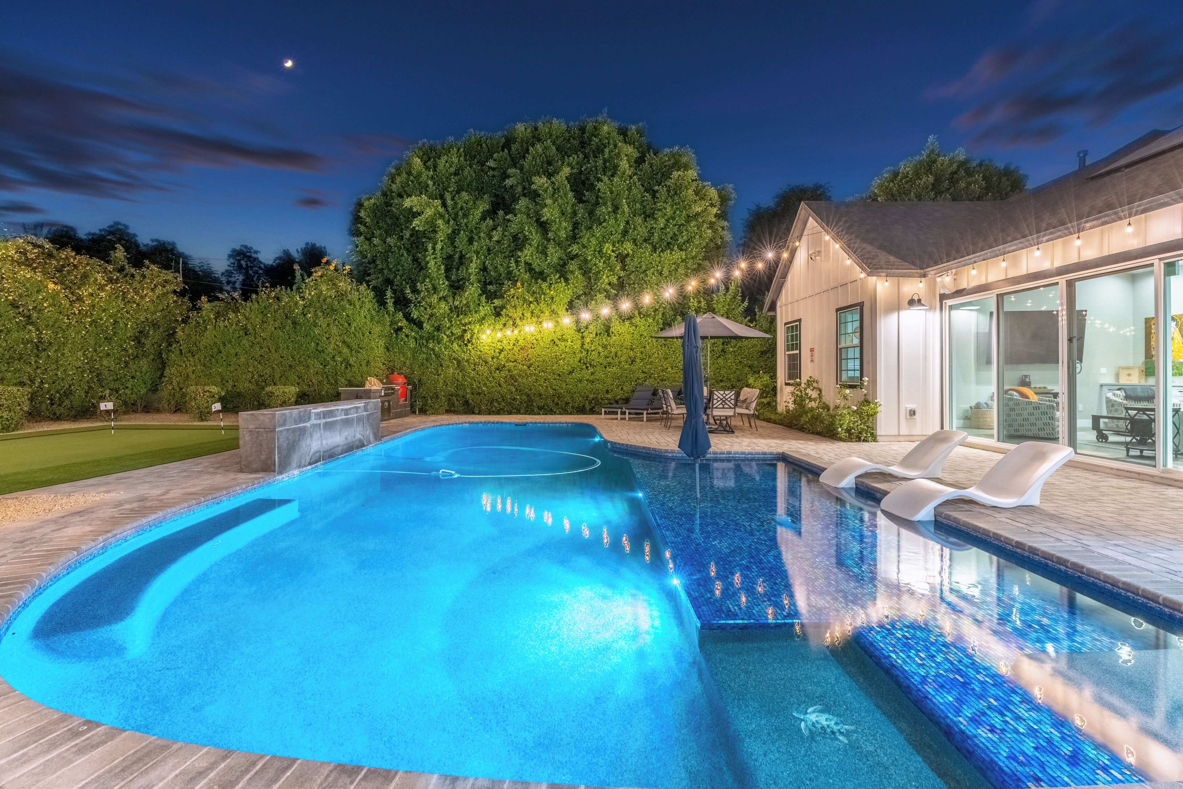 A swimming pool with a deck, a covered seating area, and a house with large windows.
