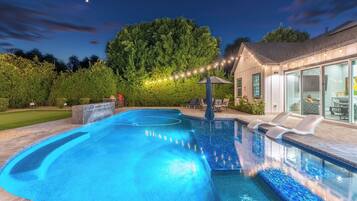 A swimming pool with a deck, a covered seating area, and a house with large windows.