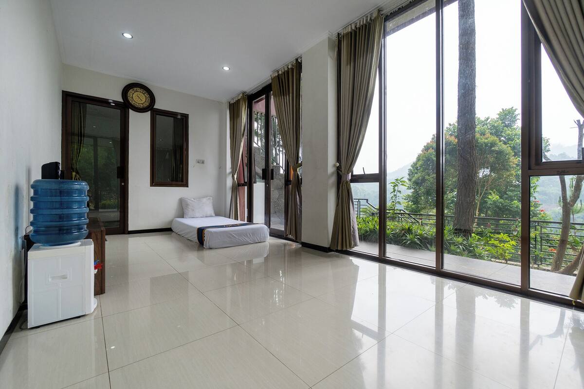 A spacious room with a bed, a water dispenser, a clock, and large windows offering a view of greenery.