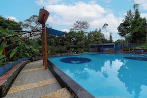 Una piscina con borde de azulejos azules y blancos, rodeada de follaje y una zona cubierta con techo azul.